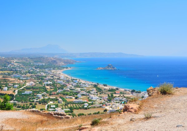 Panoramablick auf eine griechische Küstenlandschaft mit klarem, tiefblauem Meer. Im Vordergrund liegt ein trockenes, leicht hügeliges Gelände mit Felsen und Sträuchern. Dahinter erstreckt sich ein Ort mit weißen Häusern entlang eines langen Sandstrands. D