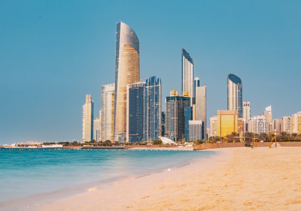 Sonnenuntergang über der Skyline von Abu Dhabi mit goldfarbenen Hochhäusern, türkisblauem Meer und einem Sandstrand im Vordergrund