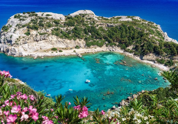 Panoramablick auf eine türkisfarbene Bucht mit felsiger Küste, dicht bewachsen mit Kiefern und Sträuchern. Im Vordergrund blühen pinke und weiße Oleanderblüten. Die Bucht liegt geschützt zwischen hellen Felsvorsprüngen und wird von mehreren kleinen Booten