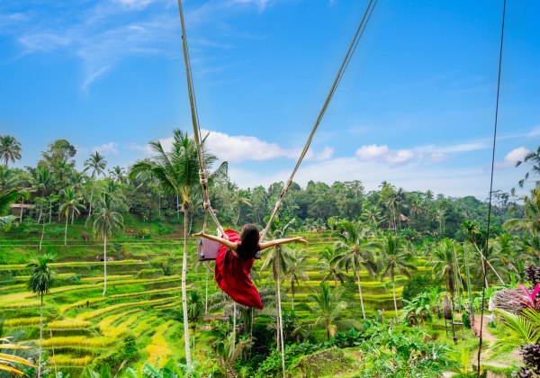Eine Frau in einem roten Kleid schwingt hoch über üppig grünen Reisterrassen auf einer berühmten Dschungelschaukel auf Bali. Sie hat die Arme weit ausgebreitet und blickt in die tropische Landschaft mit zahlreichen Palmen. Der Himmel ist blau mit wenigen 