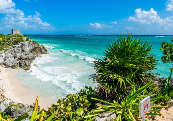 Panoramaansicht der Ruinen von Tulum auf einer Klippe mit tropischer Vegetation, türkisblauem Meer und Wellen, die gegen den weißen Sandstrand schlagen