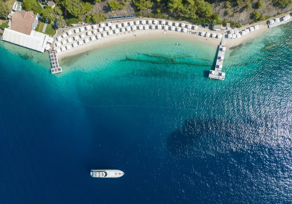 Luftaufnahme eines luxuriösen Strandabschnitts mit weißen Cabanas entlang eines hellen Sandstrands. Zwei Stege führen ins türkisfarbene Wasser, eine Yacht liegt weiter draußen im tiefblauen Meer vor Anker. Im Hintergrund dichter Pinienwald.