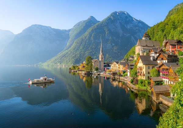 Malerisches Alpenpanorama mit dem österreichischen Ort Hallstatt am Ufer eines ruhigen Sees. Links fährt ein Ausflugsboot mit österreichischer Flagge über das spiegelglatte Wasser. Rechts am Ufer reihen sich bunte Häuser mit Holzbalkonen und Blumenschmuck