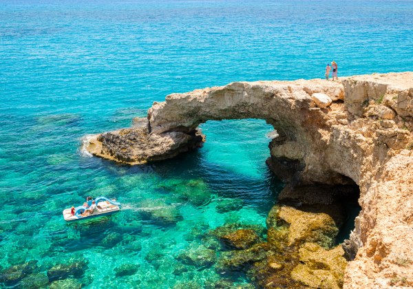 Natürlicher Felsbogen über türkisblauem Meer, ein Tretboot im Wasser und zwei Personen stehen auf dem Felsen