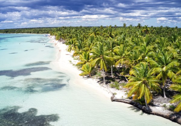 Langer weißer Sandstrand mit seichtem, türkisfarbenem Wasser. Dichter Palmenwald erstreckt sich entlang der Küste, einzelne Menschen spazieren am Strand. Im Hintergrund dramatischer Himmel mit dunklen Wolken über dem hellen Meer.