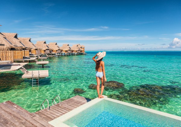 Eine Frau in einem gestreiften Badeanzug und Sonnenhut steht am Rand eines privaten Infinity-Pools und blickt auf türkisblaues, kristallklares Meer. Vor ihr befinden sich luxuriöse Wasserbungalows mit Strohdächern, die auf Stelzen über dem Wasser gebaut s