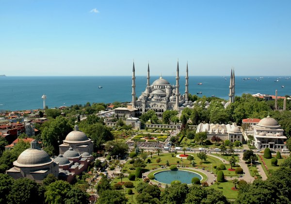 Blick auf die Blaue Moschee in Istanbul mit sechs Minaretten, umgeben von gepflegten Gärten und Springbrunnen, im Hintergrund das Meer mit zahlreichen Schiffen