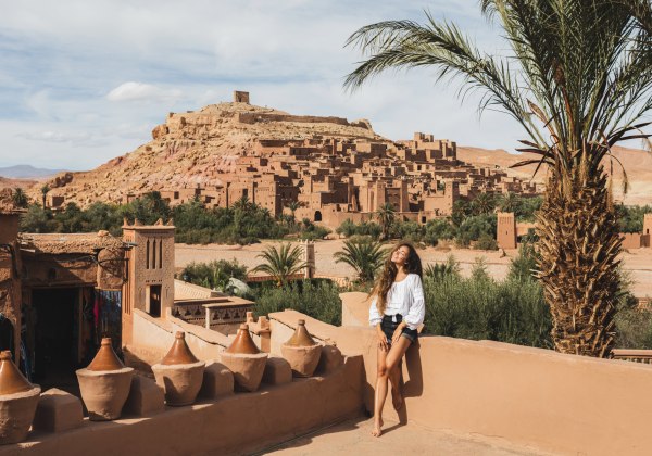 Frau lehnt entspannt an einer Mauer mit Blick auf die historische Lehmstadt Aït-Ben-Haddou in Marokko. Umgeben von Palmen, traditionellen Gebäuden und rötlich-gelben Felsen in der Wüstenlandschaft.