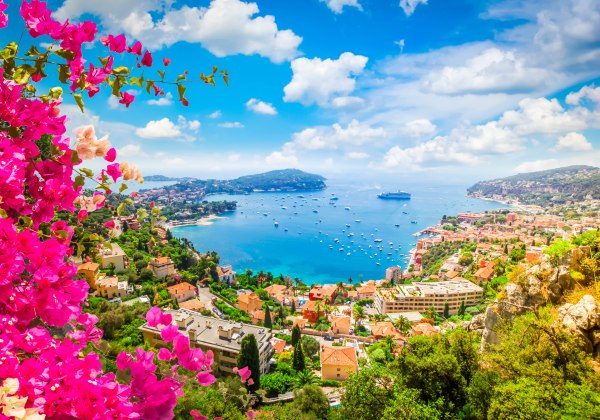 Panorama einer mediterranen Bucht mit pastellfarbenen Häusern, Bougainvillea im Vordergrund und zahlreichen Booten bis zum Horizont.