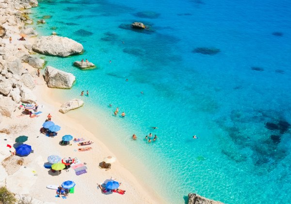Menschen baden und entspannen an einem felsigen Strand mit türkisblauem Wasser und Sonnenschirmen auf hellem Sand