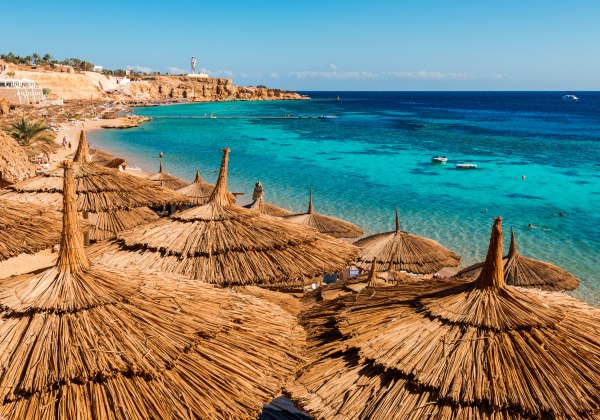Blick auf eine Bucht mit türkisfarbenem Wasser in Sharm el Sheikh. Im Vordergrund zahlreiche große Strohdach-Sonnenschirme, die sich terrassenförmig über den Hang erstrecken. Unten am Strand baden Menschen im Meer, während kleine Boote auf dem Wasser trei