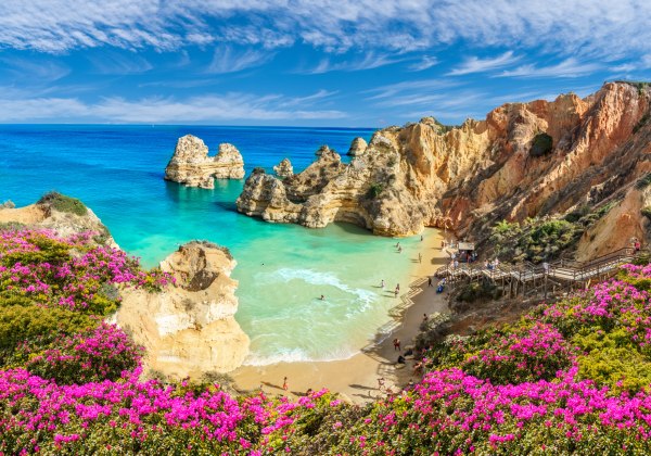 Felsige Küste der Algarve mit türkisblauem Wasser, rosa Blumen und Sandstrand zwischen Klippen