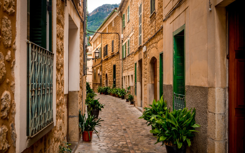 Mallorca - Sóller Eine malerische Altstadtstraße auf Mallorca, gesäumt von traditionellen Steinhäusern mit grünen Fensterläden und üppigen Topfpflanzen. Kopfsteinpflaster bedeckt den Boden, und im Hintergrund sind Berge zu sehen. Die Straße wirkt ruhig und einladend.