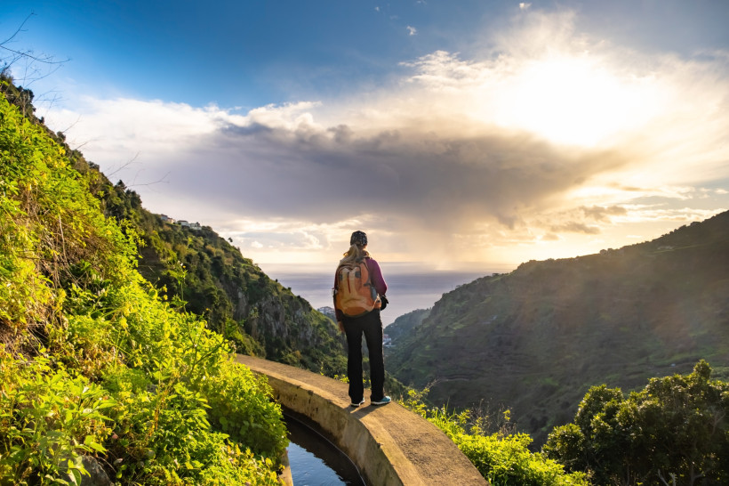 Madeira  Eine Frau mit Rucksack steht auf einem Levada-Weg auf Madeira und blickt über ein grünes Tal in Richtung Meer. Die Sonne bricht dramatisch durch die Wolken und beleuchtet die hügelige Landschaft mit warmem Licht.