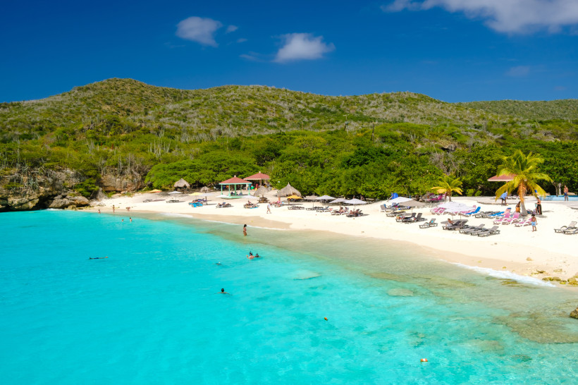 Curaçao - Grote Knip Breiter Sandstrand auf Curaçao mit zahlreichen Sonnenliegen und Sonnenschirmen vor türkisblauem Meer. Einige Menschen baden oder entspannen am Strand. Im Hintergrund grüne Hügel mit dichter Vegetation unter tiefblauem Himmel