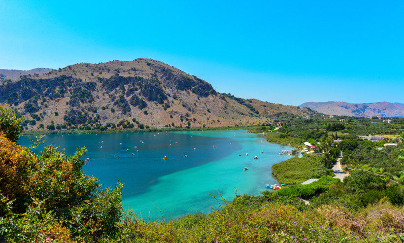 Blick über den Kournas-See mit türkisblauem Wasser, kleinen Booten und Hügeln im Hintergrund