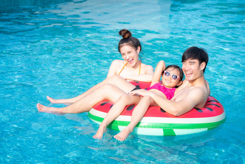 Thailand Pattaya Lachende Familie entspannt auf einer Wassermelonen-Luftmatratze im Pool. Das Kind trägt Sonnenbrille und liegt zwischen den Eltern, alle genießen sichtlich den Moment im Wasser