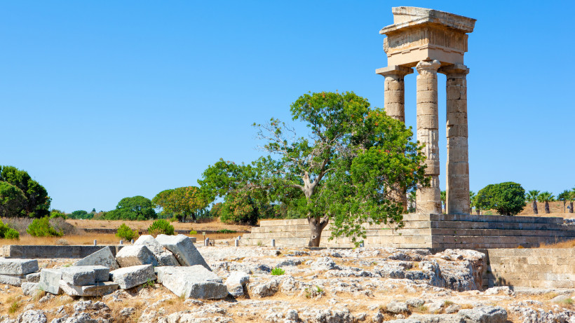 Antike Tempelruine mit vier steinernen Säulen, einem Baum und verstreuten Steinblöcken in einer archäologischen Stätte unter blauem Himmel