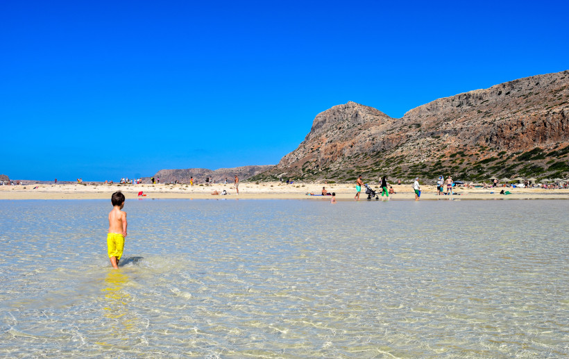 Kind steht im seichten Wasser der Bucht von Balos, im Hintergrund Strand und Felsen