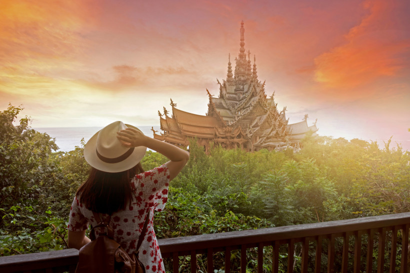 Thailand Pattaya Frau mit Hut und Rucksack steht auf einem Balkon und blickt auf das kunstvoll geschnitzte Holzgebäude Sanctuary of Truth in Pattaya bei Sonnenuntergang. Im Vordergrund dichter grüner Wald, dahinter das Meer im Abendlicht