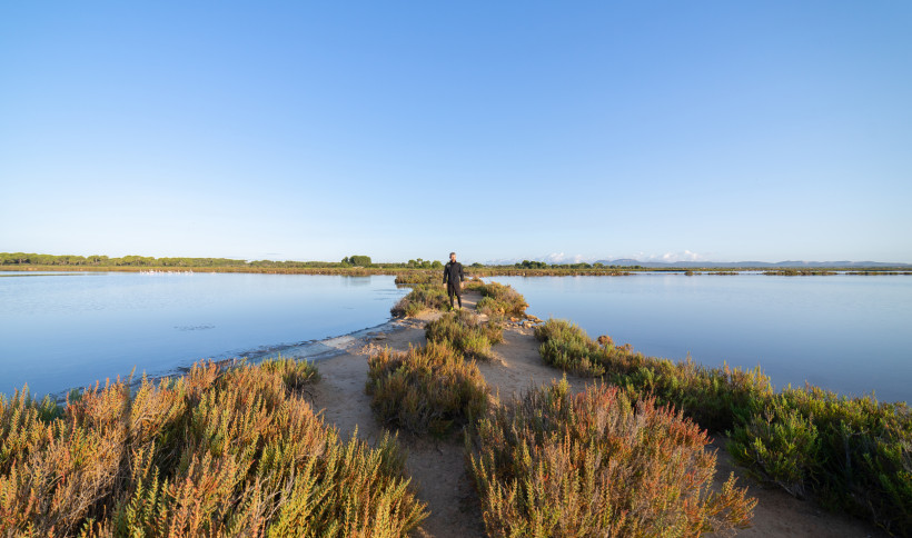 Salobrar de Campos: schmaler Pfad durch Küstenvegetation zwischen zwei flachen Lagunen, Person in der Ferne, klarer Himmel
