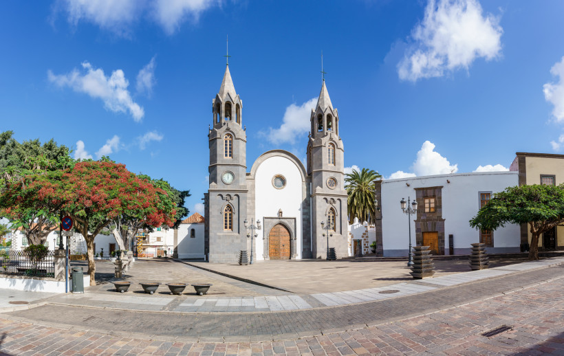 Historischer Dorfplatz mit Kirche in Telde – Gran Canaria entdecken Kirche San Juan Bautista auf dem zentralen Platz von Telde, Gran Canaria – historischer Ortskern mit Palmen, Lavastein-Fassade und blauem Himmel.
