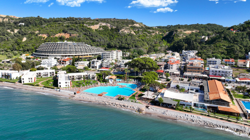 Drohnenaufnahme eines Urlaubsortes auf Rhodos mit Strandpromenade, großer Poolanlage, Hotels, türkisblauem Meer und bewaldeten Hügeln