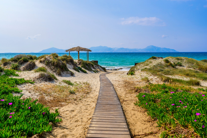 Kos Ein Holzsteg führt durch eine Dünenlandschaft mit grünem Bewuchs und rosa Blüten zu einem türkisblauen Meer. Im Hintergrund sind Berge und eine strohgedeckte Schattenspender-Struktur zu sehen. Der Himmel ist hellblau mit wenigen Wolken.