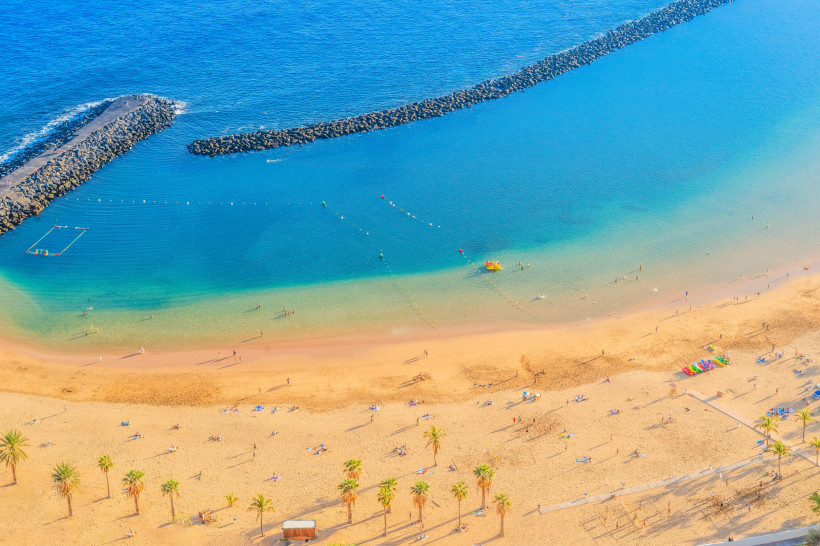 Playa de Las Teresitas – goldener Traumstrand bei Santa Cruz Luftaufnahme des Playa de Las Teresitas auf Teneriffa mit einem breiten goldgelben Sandstrand, gesäumt von Palmen. Viele Menschen liegen am Strand oder schwimmen im klaren, türkisblauen Wasser. Zwei lange Wellenbrecher schützen die ruhige Bucht, sodass da
