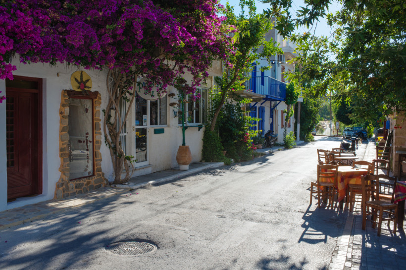 Schmale mediterrane Dorfstraße mit weißen Häusern, üppig blühender Bougainvillea, kleinen Geschäften und einem Straßencafé mit Holztischen und Stühlen im Schatten
