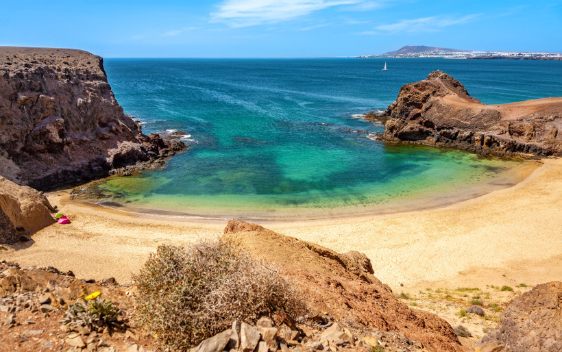 Lanzarote Kleine, abgelegene Sandbucht mit türkisfarbenem Wasser, umgeben von Felsen. Am Strand liegen zwei bunte Sonnenschirme, im Hintergrund ist das offene Meer zu sehen.