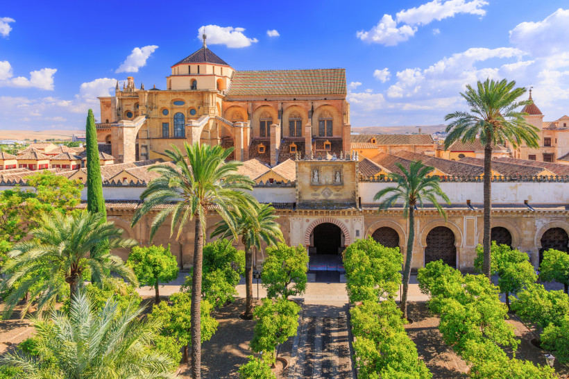 Blick auf die Mezquita-Kathedrale von Córdoba mit maurisch-christlicher Architektur. Im Vordergrund ein grüner Orangenhof mit Palmen, im Hintergrund das imposante Gebäude mit Bögen, Zinnen und Türmen unter blauem Himmel mit weißen Wolken.