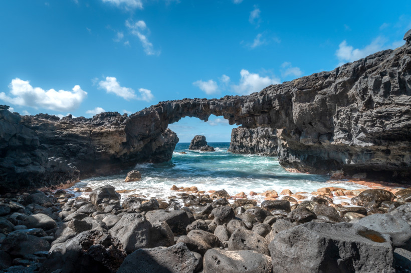 Felsiger Strand mit einem natürlichen Felsbogen in Las Puntas. Insel El Hierro. Kanarische Inseln. Natursteinbrücke aus dunklem Lavagestein über dem Meer, mit rauer Küste und Felsen im Vordergrund bei sonnigem Wetter