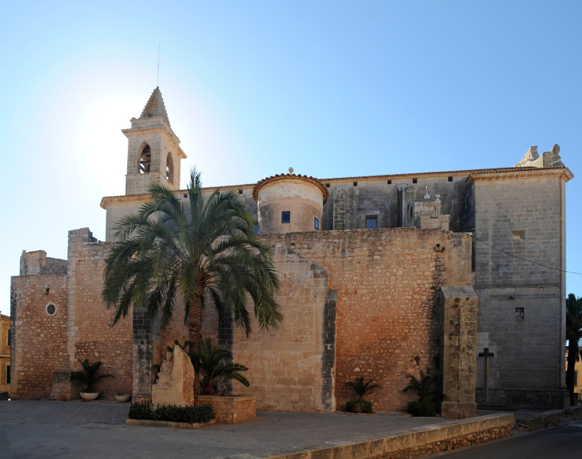 Mallorca - Santanyí Historische Kirche aus Naturstein mit einem Glockenturm und einer kleinen Kuppel auf dem Dach. Die Kirche hat eine Mischung aus rötlich-braunem und grauem Steinmauerwerk. Im Vordergrund steht eine große Palme und eine kleine Statue, die von niedrigen Pfla