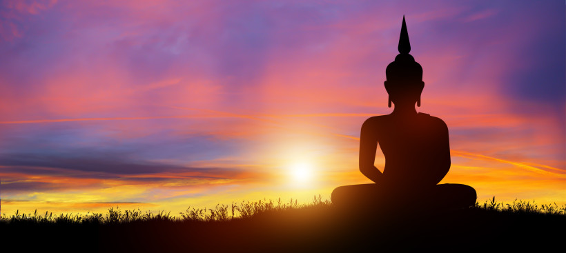 Thailand Pattaya Silhouette einer Buddha-Statue im Lotussitz vor farbenprächtigem Sonnenuntergang mit orange-lila Himmel. Im Vordergrund Gras und Pflanzen, die Szene wirkt friedlich und meditativ.