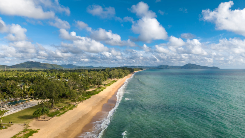 Luftaufnahme von Mai Khao Beach mit langem Sandstrand, Küstenlinie, türkisfarbenem Meer und bewaldetem Hinterland unter wolkigem Himmel