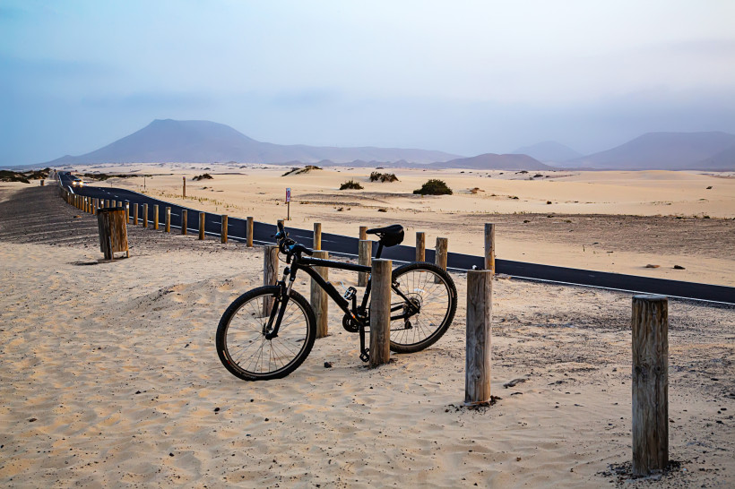 Fahrrad an Holzpfosten neben Asphaltstraße in den Sanddünen bei Corralejo, im Hintergrund Berge