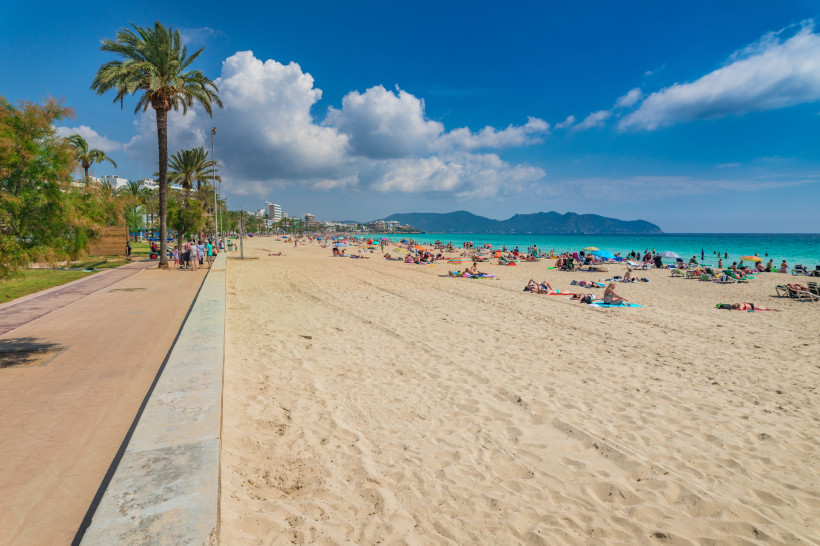 Großer Sandstrand auf Mallorca mit Palmenpromenade, vielen Badegästen, Sonnenliegen und türkisfarbenem Meer vor bergiger Küstenlandschaft
