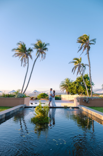 Südafrika Ein Paar steht in romantischer Pose an einem kleinen Teich, eingerahmt von Palmen, mit Blick auf ein luxuriöses Resort im Hintergrund. Die Szene spielt sich bei klarem Himmel und weichem Abendlicht ab – perfekte Urlaubsstimmung.