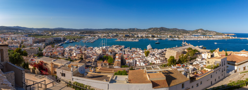Ibiza-Stadt Panorama über Ibiza-Stadt: weiße Altstadthäuser, Festungswälle, Yachthafen und tiefblaue Bucht mit Bergen im Hintergrund.