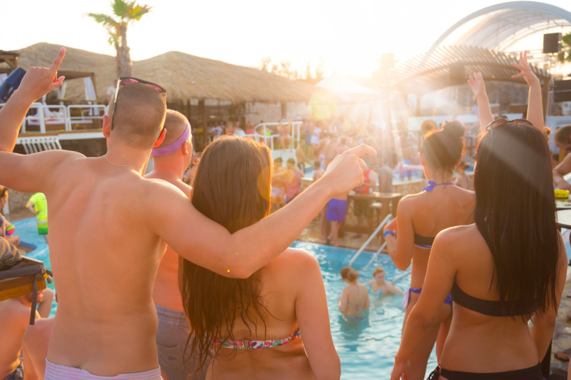 Bulgarien Menschen feiern eine lebhafte Poolparty bei Sonnenuntergang. Ein junger Mann und eine Frau in Badekleidung stehen Arm in Arm am Poolrand, während viele andere im Wasser und auf der Terrasse tanzen und feiern. Im Hintergrund sind Palmen, Strohdächer und ei