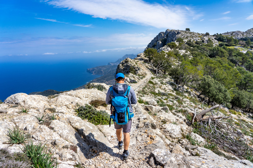 Mallorca Wanderer mit blauem Rucksack und Basecap geht auf einem felsigen Wanderweg in den Bergen von Mallorca. Der Weg schlängelt sich durch eine zerklüftete Landschaft mit grüner Vegetation und kleinen Bäumen. Im Hintergrund öffnet sich ein atemberaubender Blick