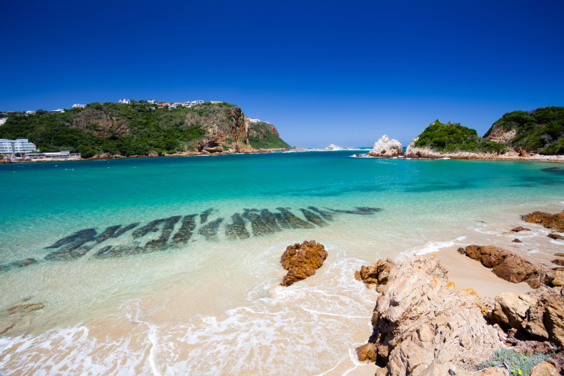 Südafrika Blick auf einen traumhaften Strand in Südafrika mit türkisblauem Wasser und feinem Sand. Im Hintergrund erheben sich grüne Klippen, die teilweise von weißen Gebäuden gesäumt sind. Der Himmel ist strahlend blau – perfektes Strandwetter.