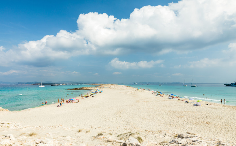 Formentera - Playa de Ses Illetes Blick auf die Playa de Ses Illetes auf Formentera mit weißem Sandstrand, Dünen und türkisgrünem Meer. Mehrere Yachten und Boote liegen vor der Küste, im Hintergrund sind Berge sichtbar.