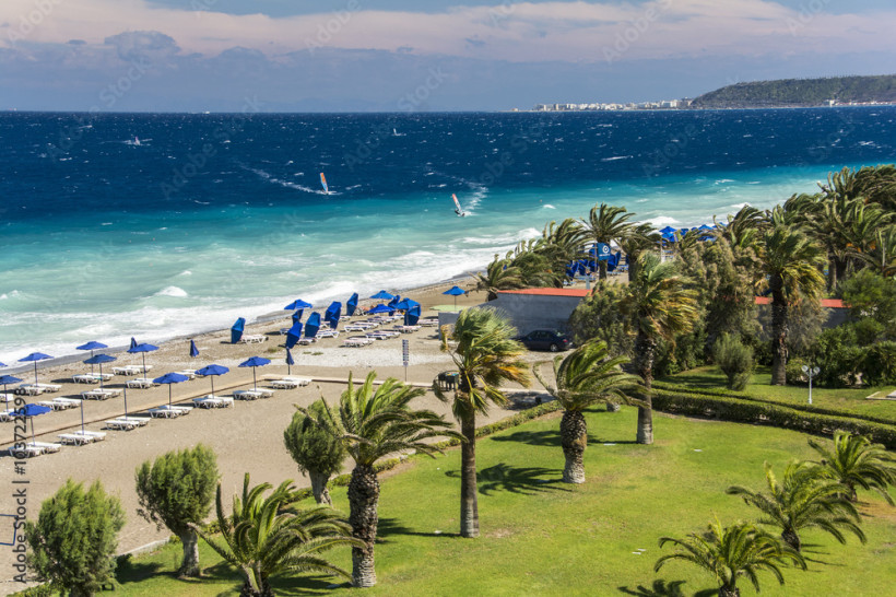 Palmenstrand auf Rhodos mit starkem Wind, blauen Sonnenschirmen, hohen Wellen und Windsurfern im türkisfarbenen Meer