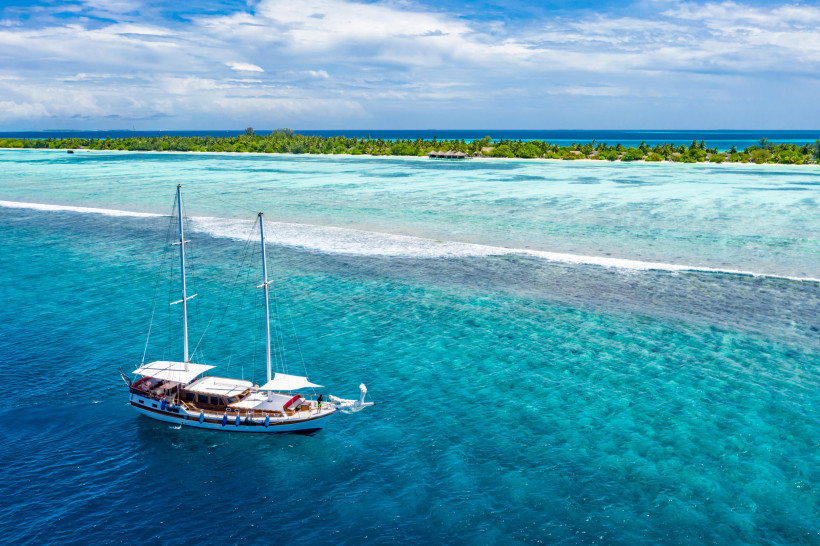 Malediven Ein elegantes Segelboot gleitet durch das türkisfarbene Wasser einer tropischen Insel. Die sanften Wellen und die üppige Vegetation im Hintergrund schaffen eine traumhafte Kulisse für ein unvergessliches Segelerlebnis.
