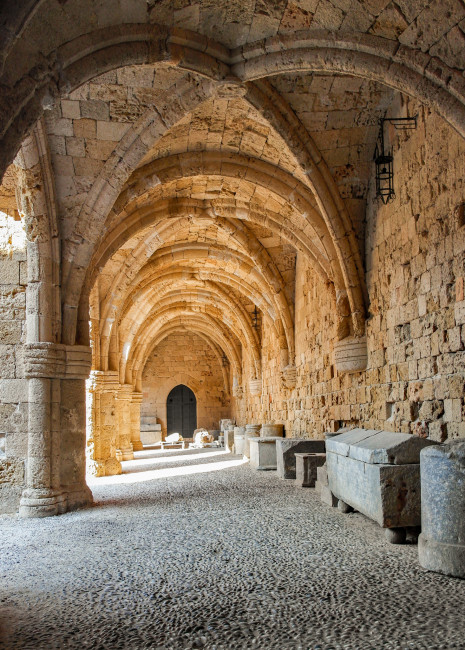  Das Bild zeigt einen mittelalterlichen Gewölbegang mit aufeinandergestapelten Spitzbögen und robustem Mauerwerk aus gelbem Naturstein.   Auf der rechten Seite des Ganges stehen mehrere steinerne Sarkophage oder Truhen entlang der Wand.   Der Boden ist mi