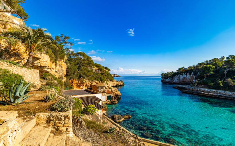 Mallorca - Cala Llombards Ein Blick auf eine kleine Küstenbucht mit kristallklarem, türkisfarbenem Wasser. Umgeben von Felsen, Agaven und mediterranen Pflanzen führt ein Weg entlang der Felsklippen. Die Landschaft wirkt naturbelassen und lädt zu einem Spaziergang ein.