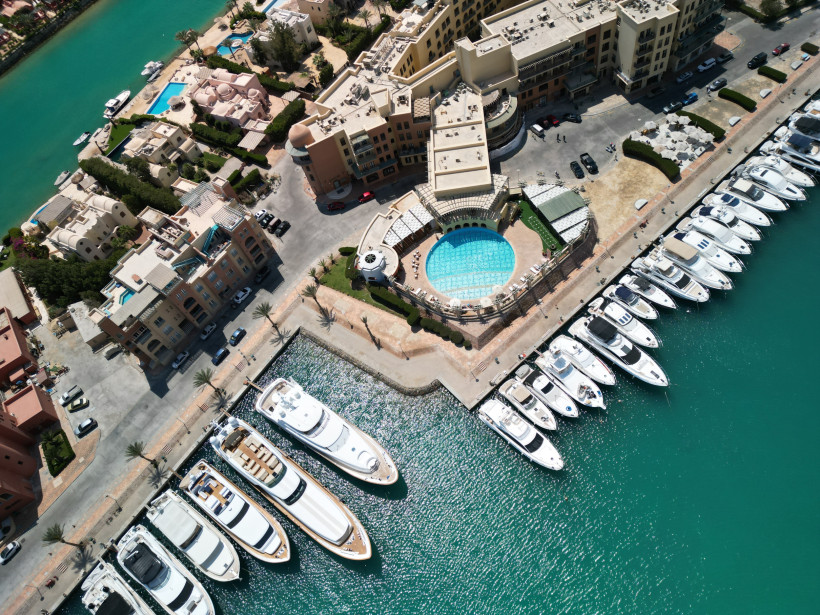 Ägypten - El Gouna Luftaufnahme einer Marina mit einem großen runden Pool, umgeben von modernen Gebäuden, Palmen und einer Reihe weißer Yachten im türkisfarbenen Wasser.