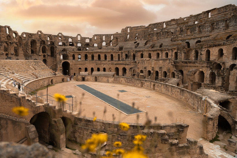 Blick ins römische Amphitheater von El Djem in Tunesien bei Sonnenuntergang. Die monumentalen Steinmauern des Kolosseums ragen in den Himmel, während im Vordergrund gelbe Blumen unscharf die Szene einrahmen. Einige Menschen schlendern durch das untere Run